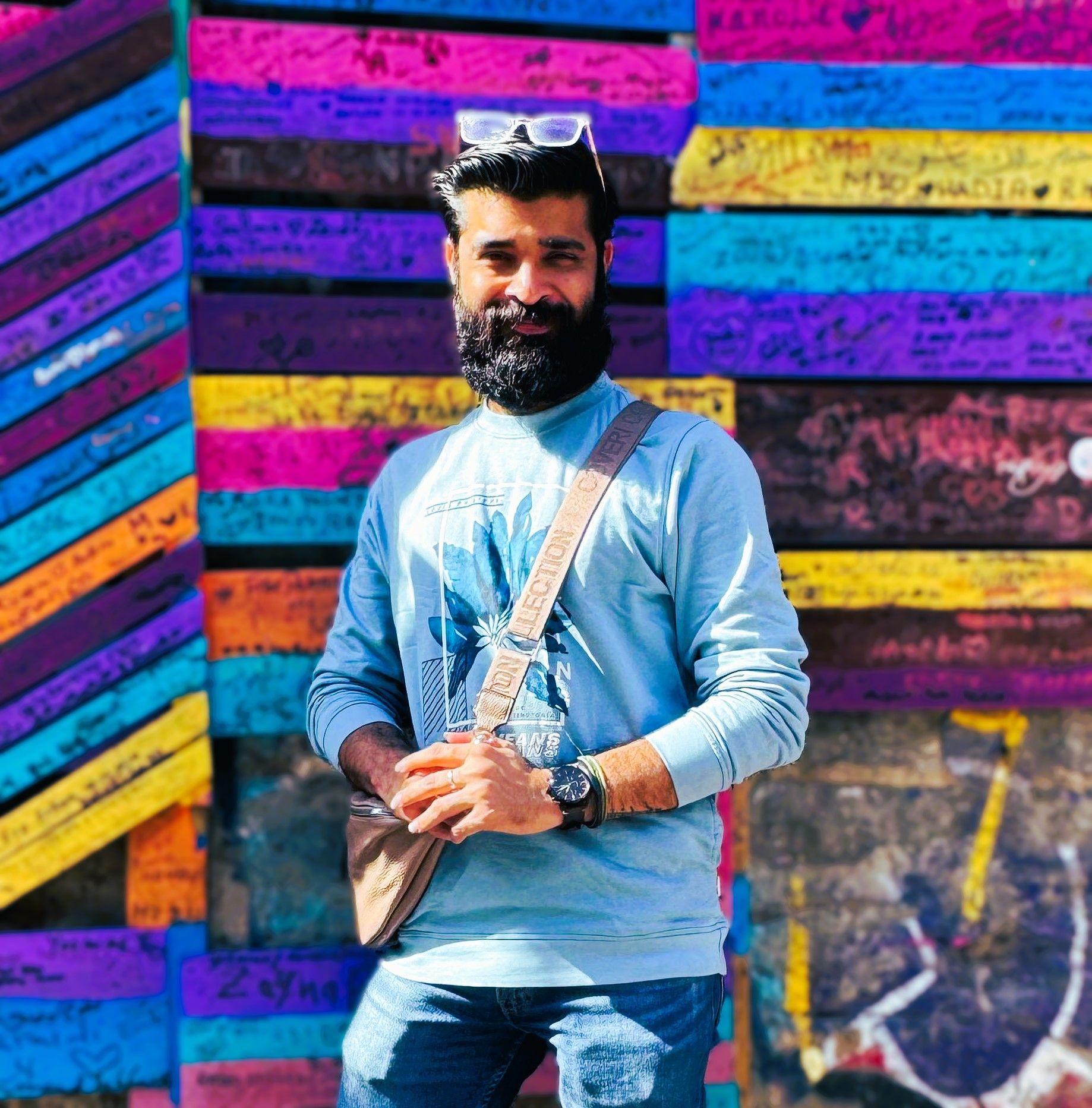 Man with beard wearing blue sweatshirt stands in front of colorful graffiti wall, holding a bag.